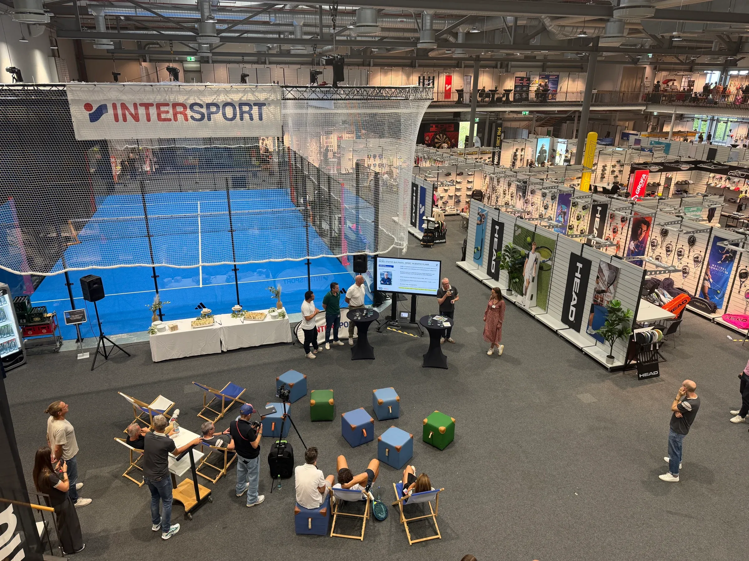 adidas PILLARFORCE FX mit blauem Turf in einer Messehalle der INTERSPORT-Messe, davor Besucher und Sitzbereiche